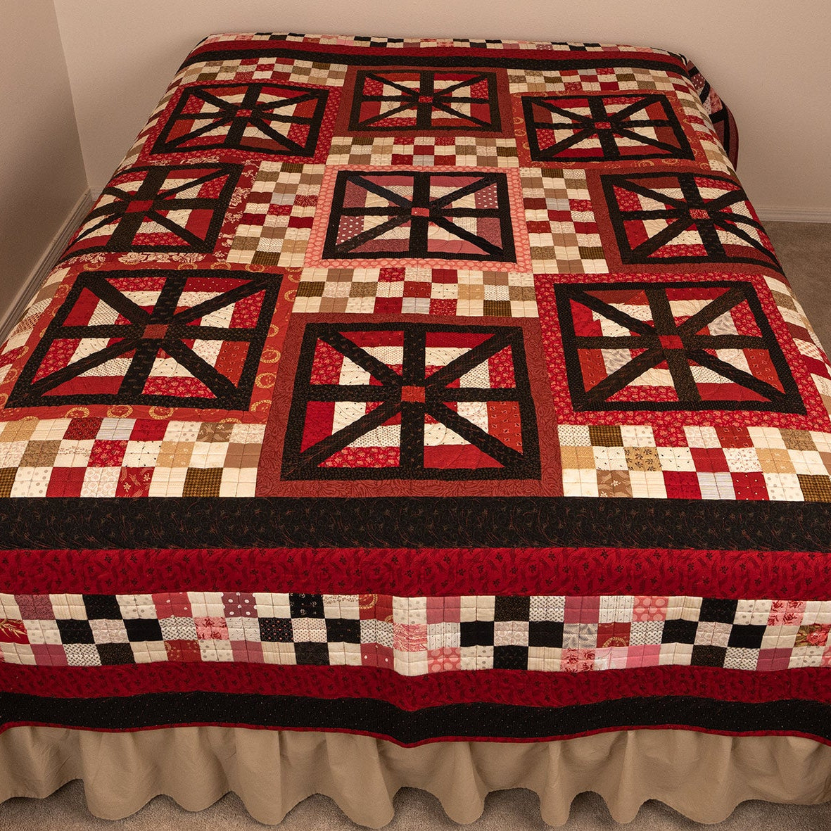 A patchwork quilt with red, black, beige, and white geometric and checkered patterns, draped over a bed