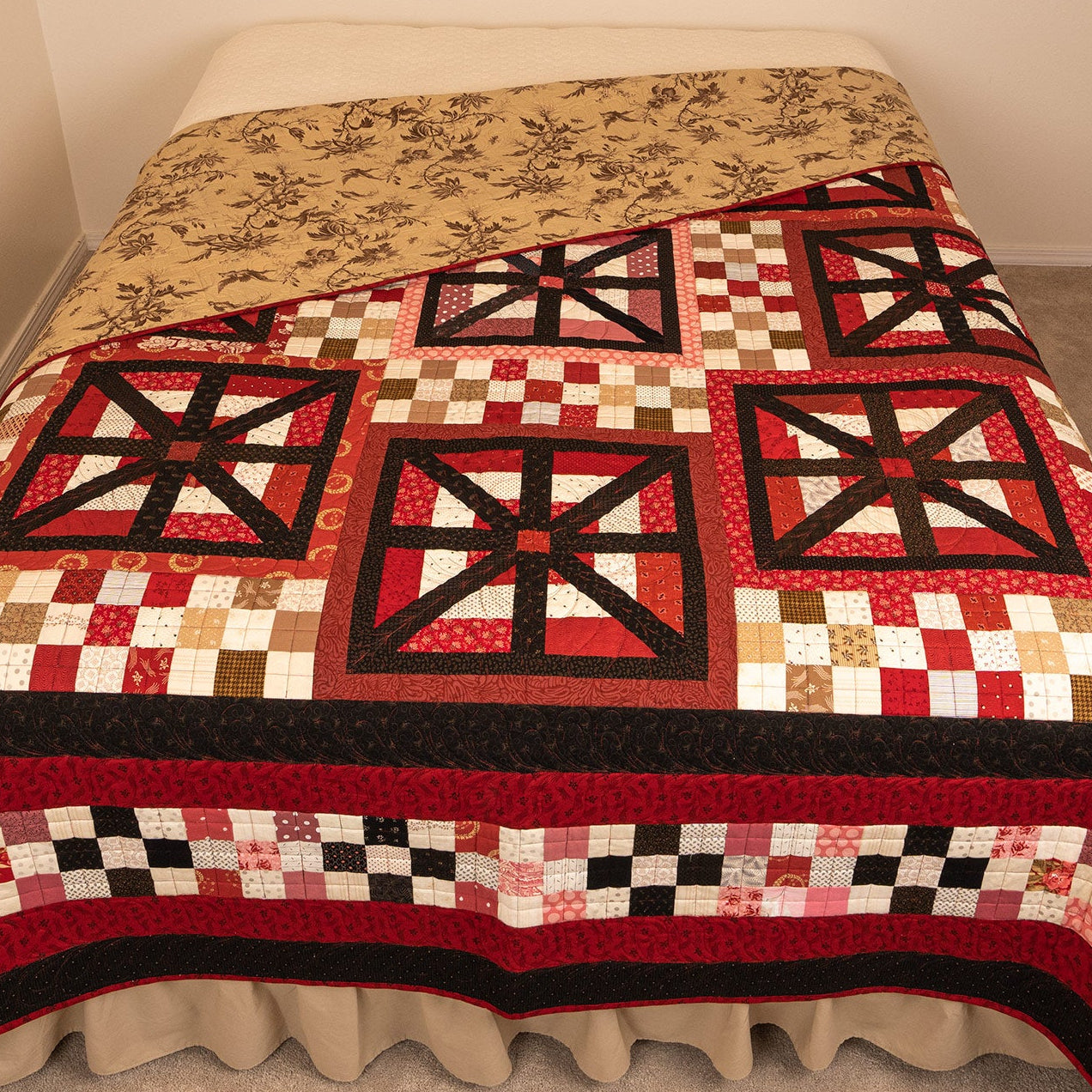 A patchwork quilt with red, black, beige, and white geometric and checkered patterns, draped over bed with the floral back being shown in a tan floral print.