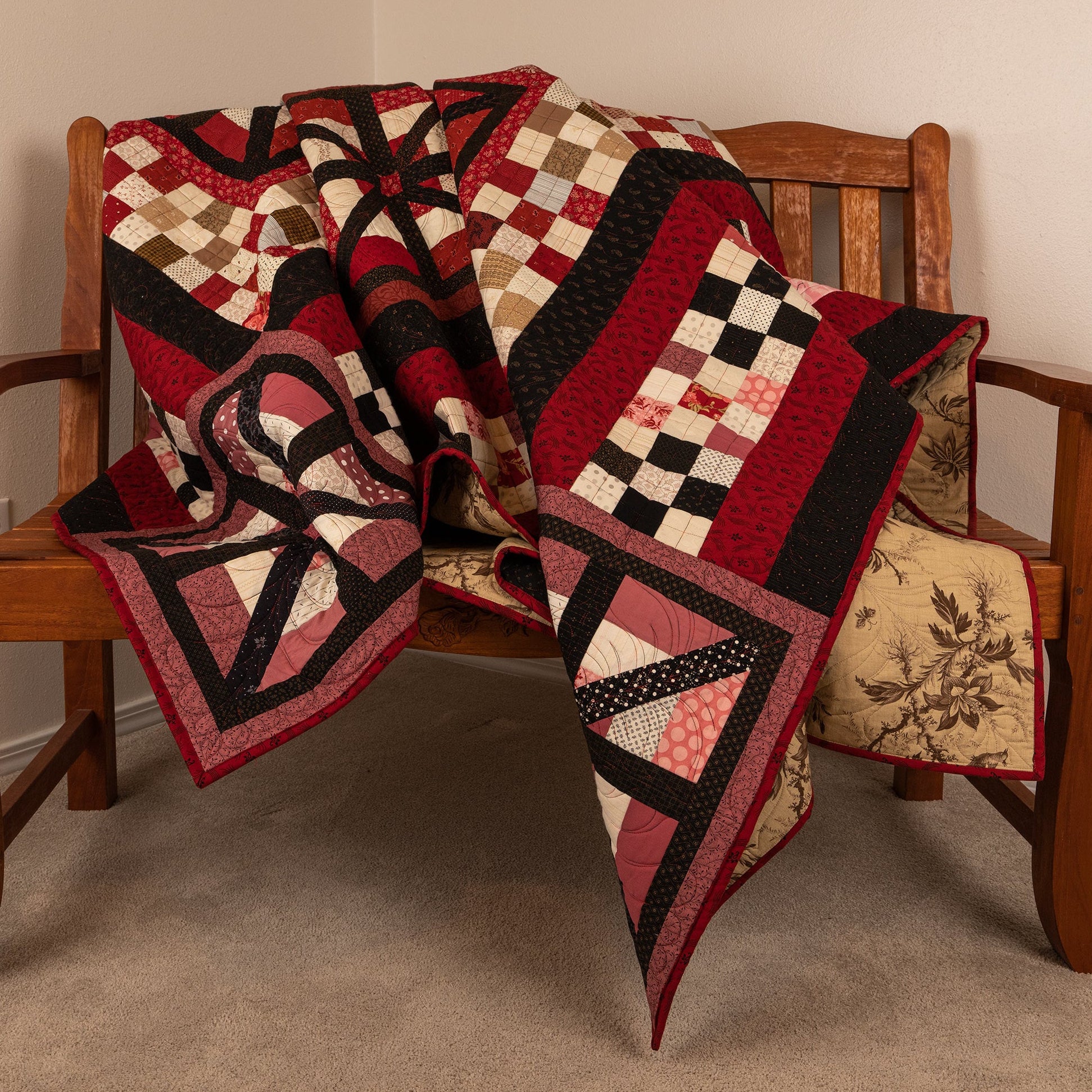 A patchwork quilt with red, black, beige, and white geometric and checkered patterns, draped over a wooden bench in a home setting.