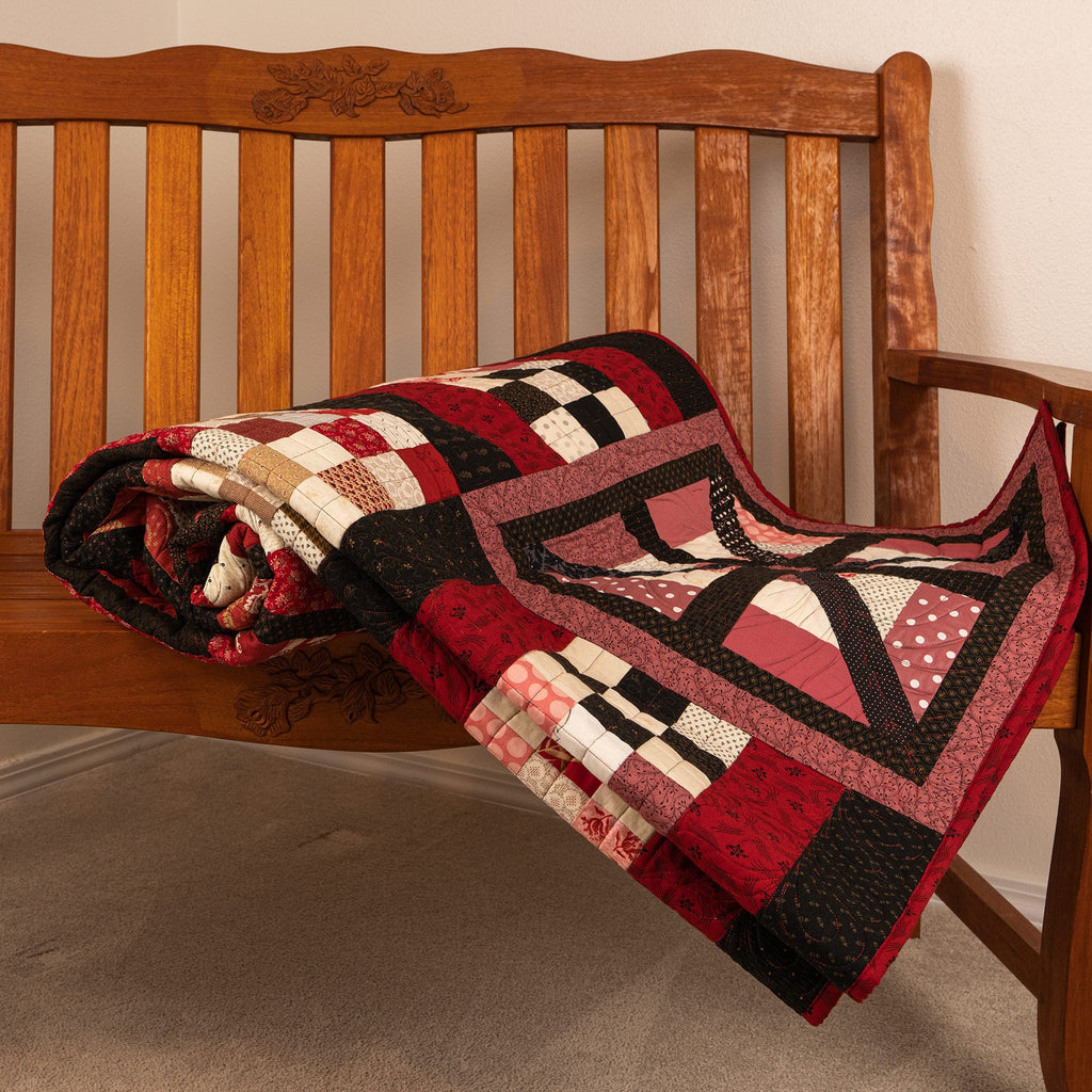 A patchwork quilt with red, black, beige, and white geometric and checkered patterns, rolled up on a wooden bench in a home setting.