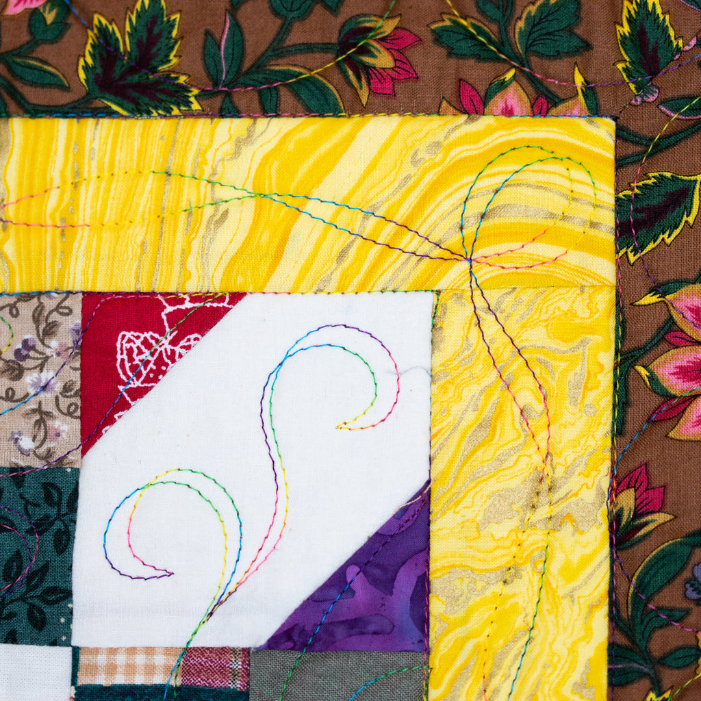 A close-up of a colorful quilted fabric panel with a patchwork design, featuring a large yellow marbled section, a white diagonal section with multicolored stitched swirls, and floral brown patterned borders.
