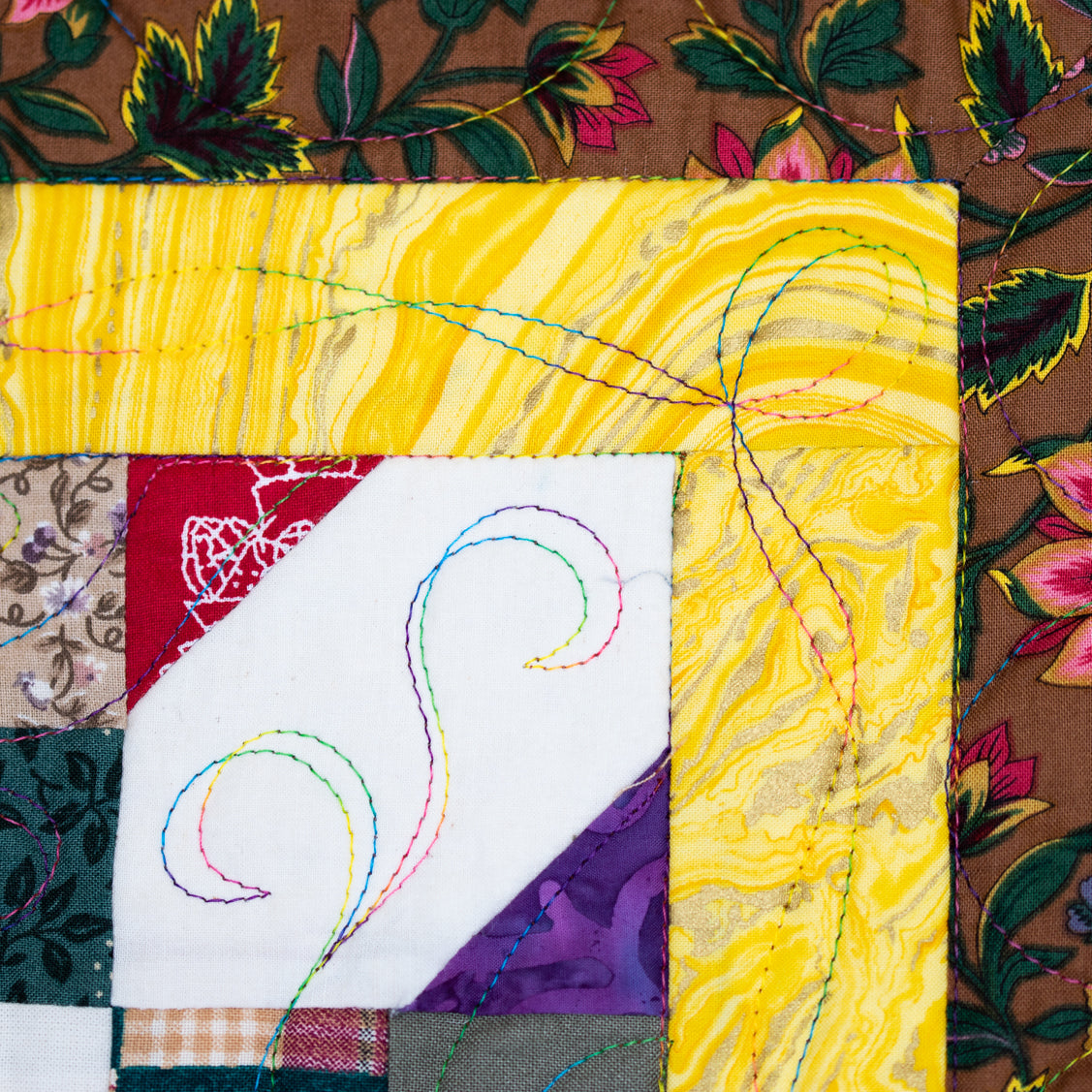 A close-up of a colorful quilted fabric panel with a patchwork design, featuring a large yellow marbled section, a white diagonal section with multicolored stitched swirls, and floral brown patterned borders.