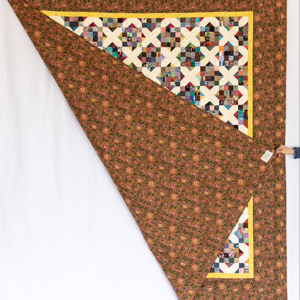 A handmade quilted blanket with a brown floral patterned body, a multicolor geometric starburst triangle, and yellow borders, shown hanging against a white backdrop.