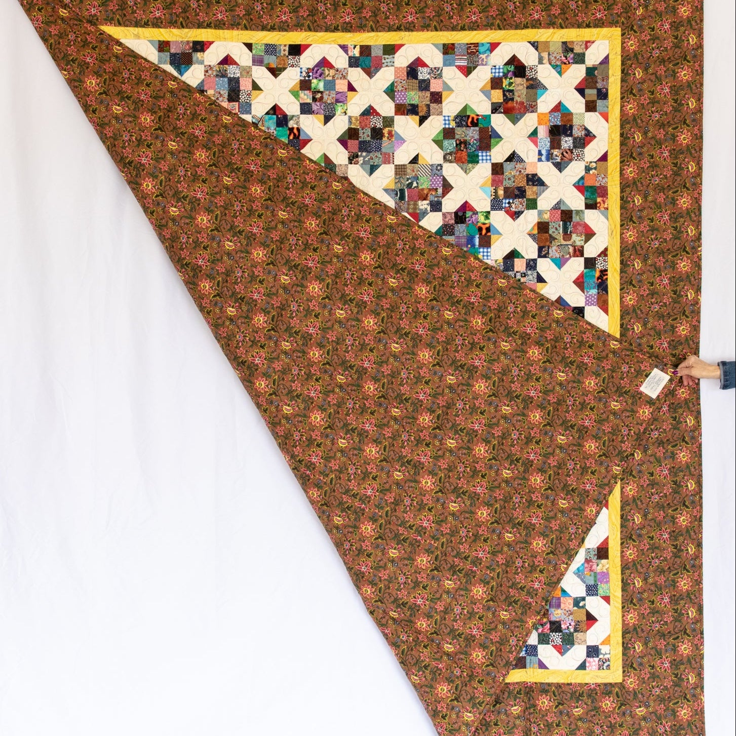 A handmade quilted blanket with a brown floral patterned body, a multicolor geometric starburst triangle, and yellow borders, shown hanging against a white backdrop.