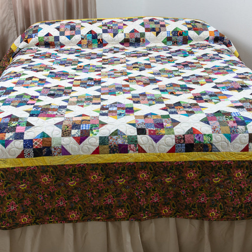 A quilted bedspread with a colorful geometric star pattern and a dark floral border, styled on a bed with a beige ruffled skirt in a bedroom setting.
