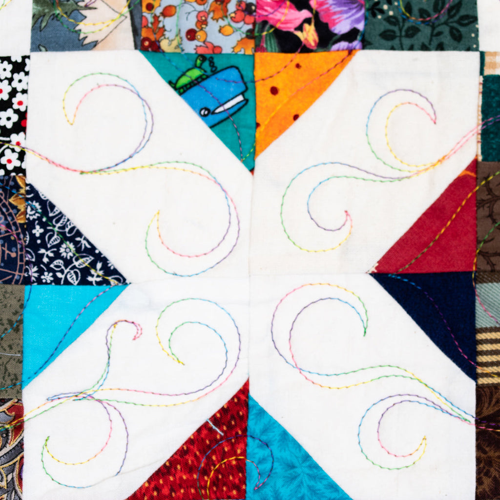 A close-up of a quilted fabric block featuring a white central square with embroidered swirl stitching, surrounded by colorful patterned fabric patches including floral, striped, and textured designs.