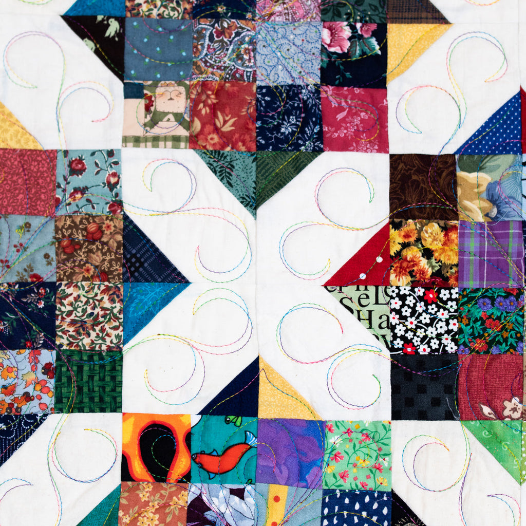 A close-up of a colorful patchwork quilt with geometric and floral fabric blocks, white quilting stitches, and a white border.