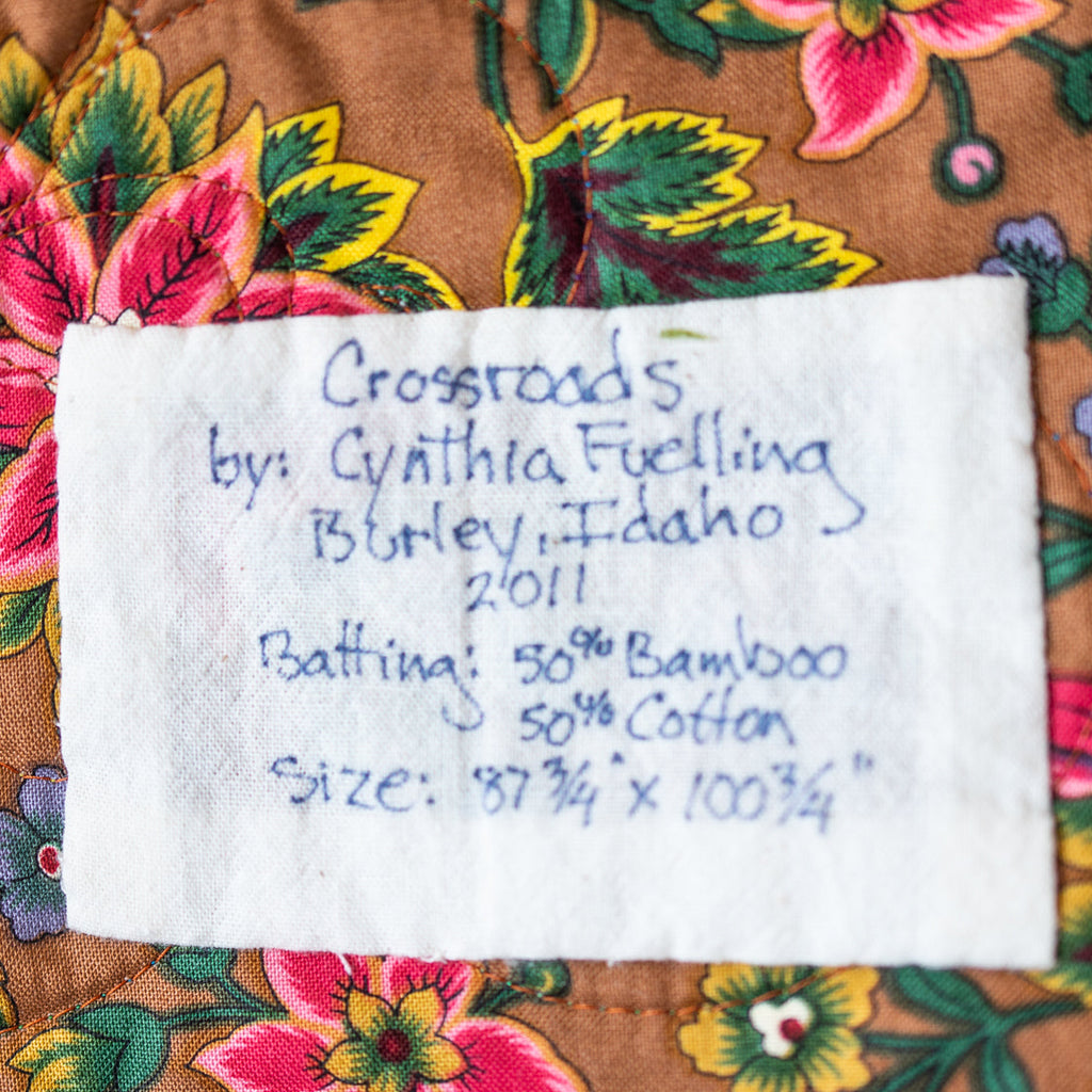 A close-up of a white fabric label sewn onto a brown floral quilted surface. The label is handwritten with the brand 'Crossroads by Cynthia Fuelling', location 'Burlley, Idaho', and batting/cotton sizing details.