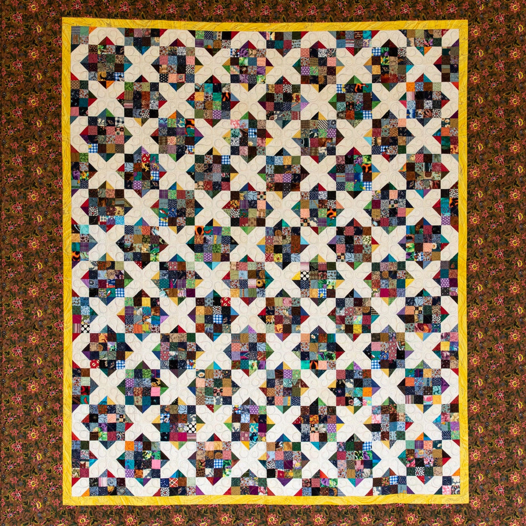 A square quilt, featuring a multicolor patchwork center with repeating cream X-shaped blocks and a wide brown floral border with a yellow inner border.