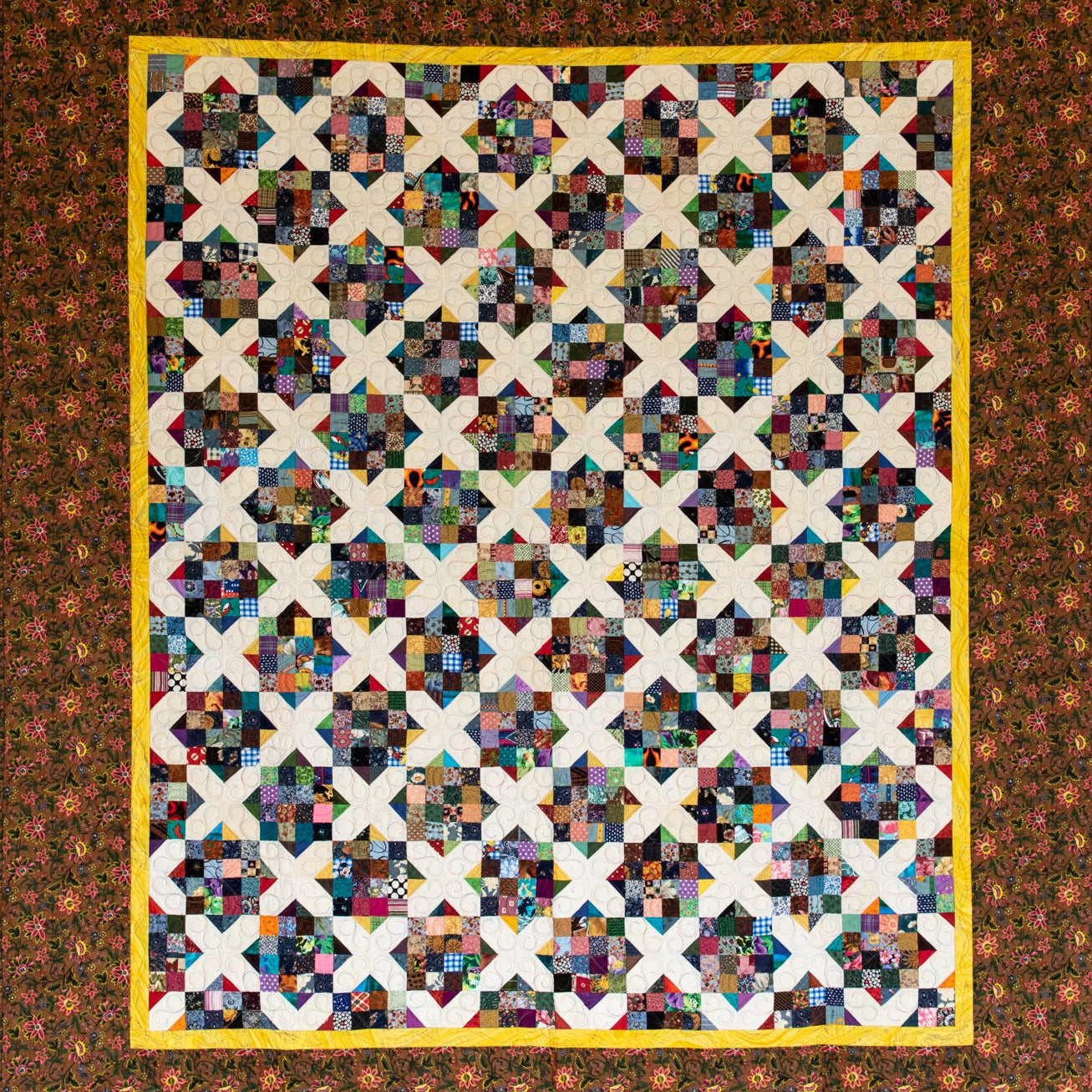 A square quilt, featuring a multicolor patchwork center with repeating cream X-shaped blocks and a wide brown floral border with a yellow inner border.