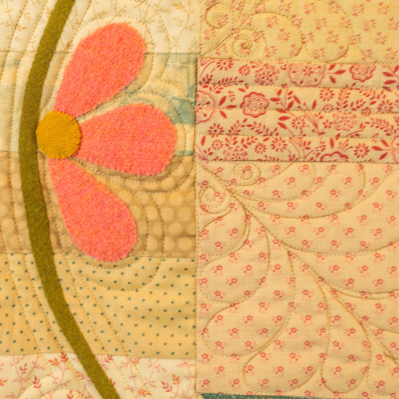A close-up of a handmade quilted textile featuring a large pink flower with a yellow center, cream and pink patterned fabric blocks, and green and teal stitched sections.
