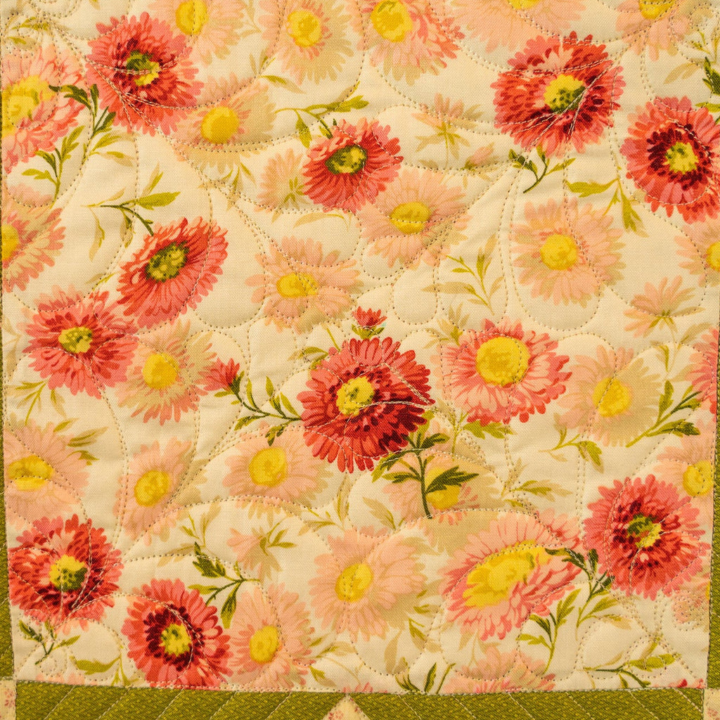 A close-up of a quilted fabric panel featuring a floral pattern with pink and red flowers, yellow centers, and green leaves on a cream background, bordered by olive green and pink patterned sections.
