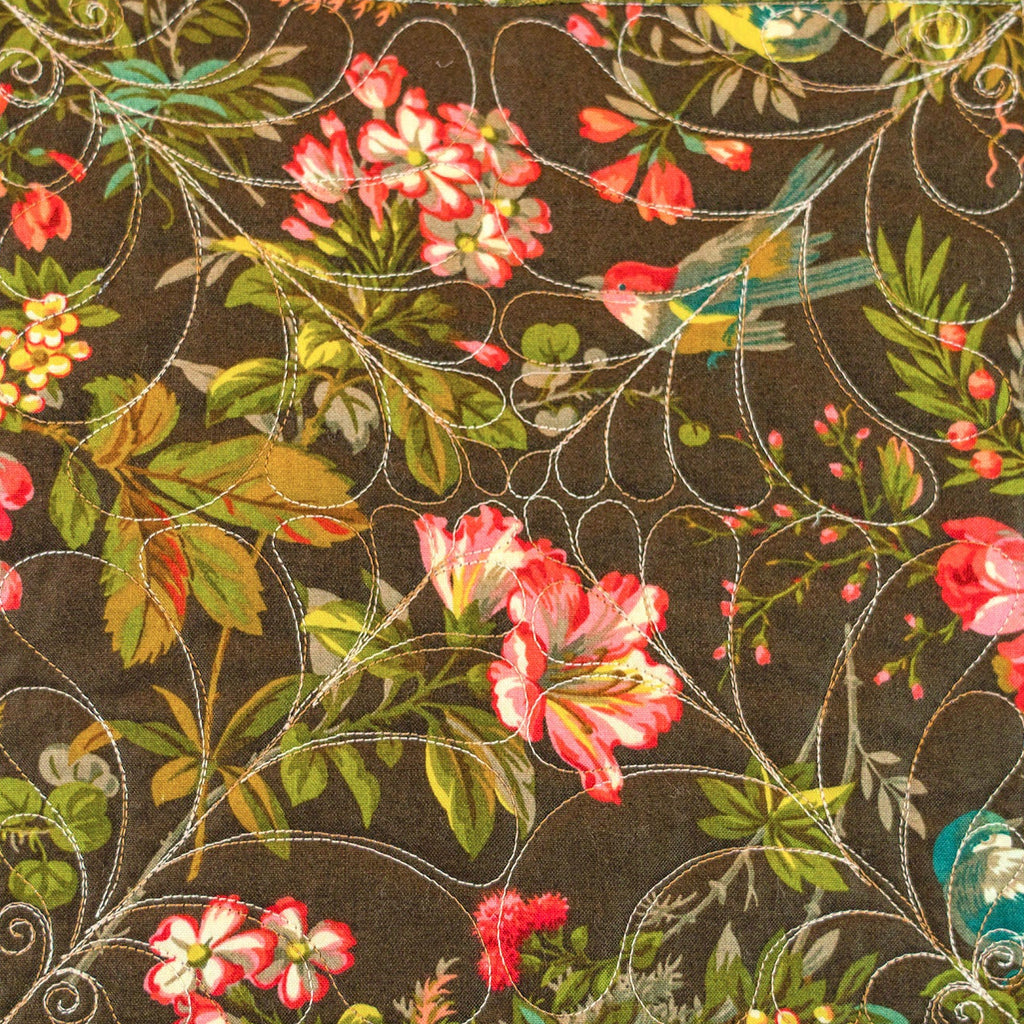 A close-up of a quilted fabric featuring a central floral and bird pattern on a dark brown background.