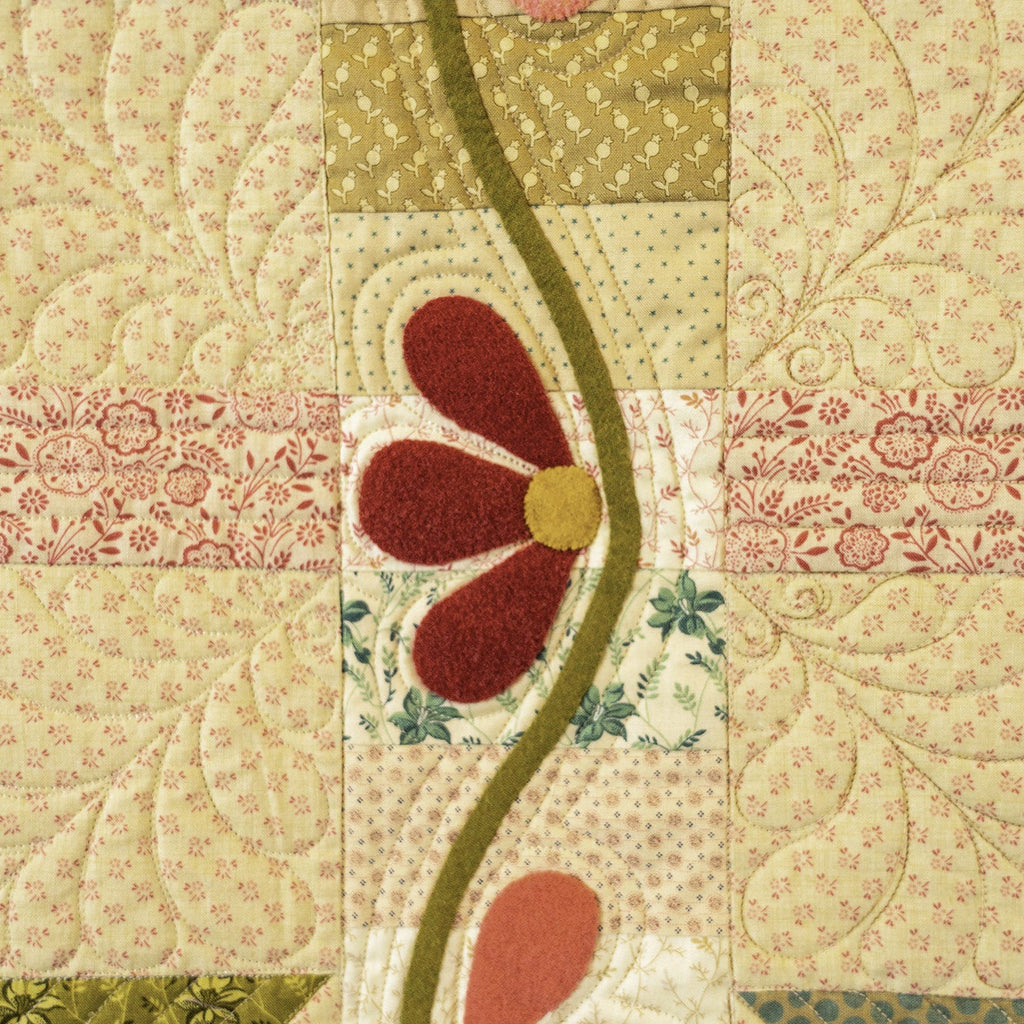 A close-up of a quilted fabric panel featuring a patchwork design with floral and geometric patterns in beige, green, pink, and red, with a central wavy olive-green stripe and a stylized flower motif.