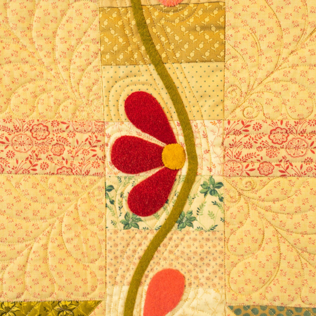 A close-up of a patchwork quilt featuring various fabric patterns in yellow, pink, green, and red, with a central red flower applique and a wavy olive-green line.