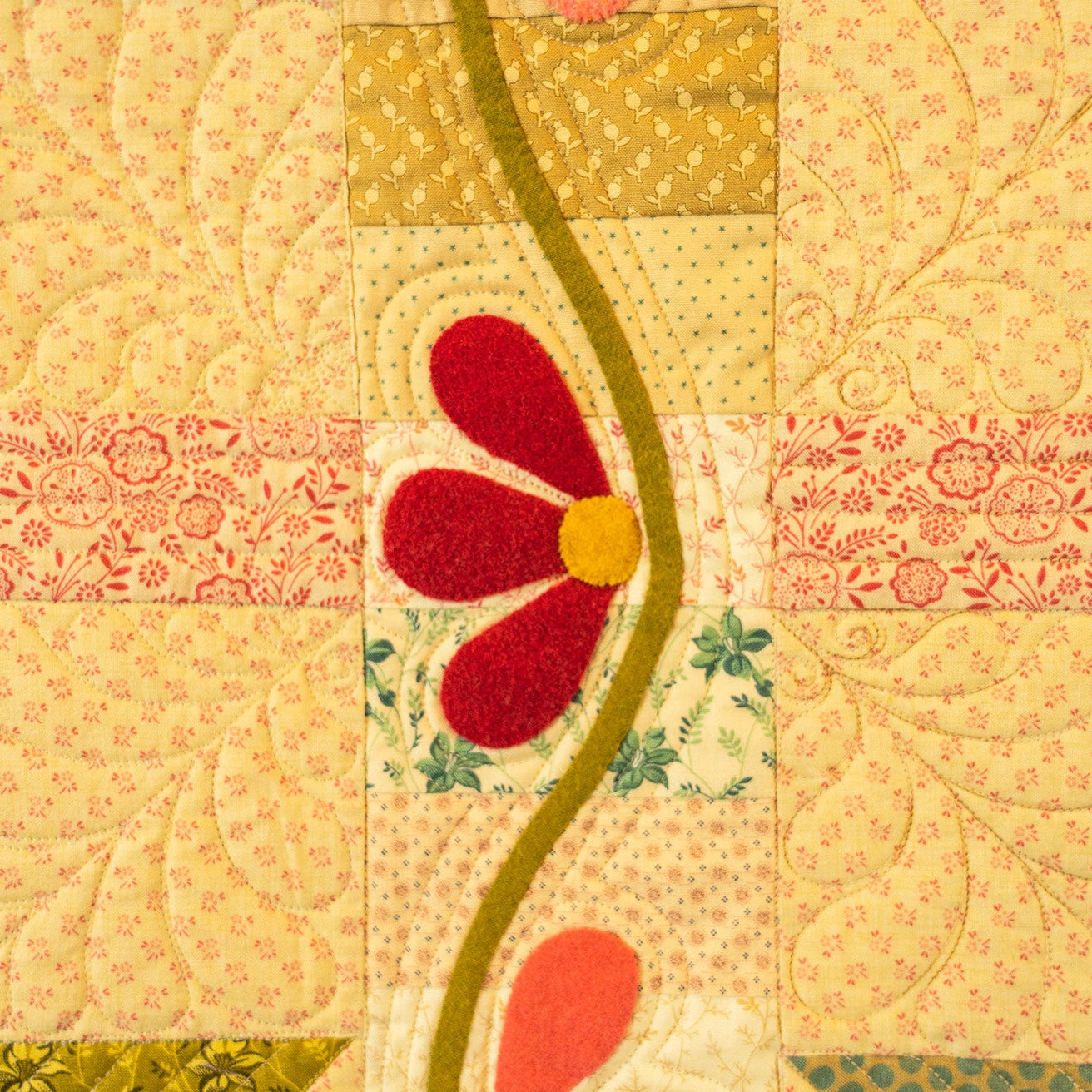 A close-up of a patchwork quilt featuring various fabric patterns in yellow, pink, green, and red, with a central red flower applique and a wavy olive-green line.