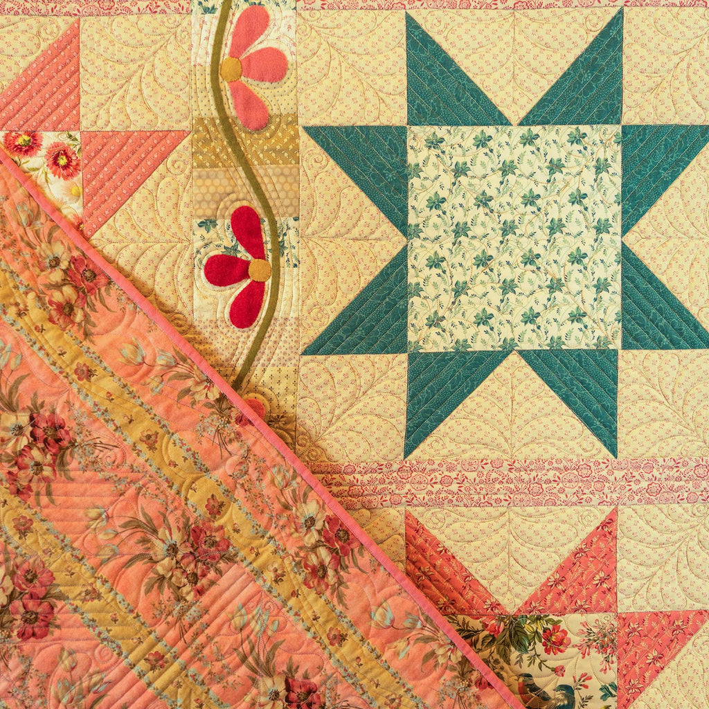 A close-up of a vintage-style quilted textile featuring a star and floral block pattern in coral, teal, cream, and pink, with a diagonal floral ribbon detail.
