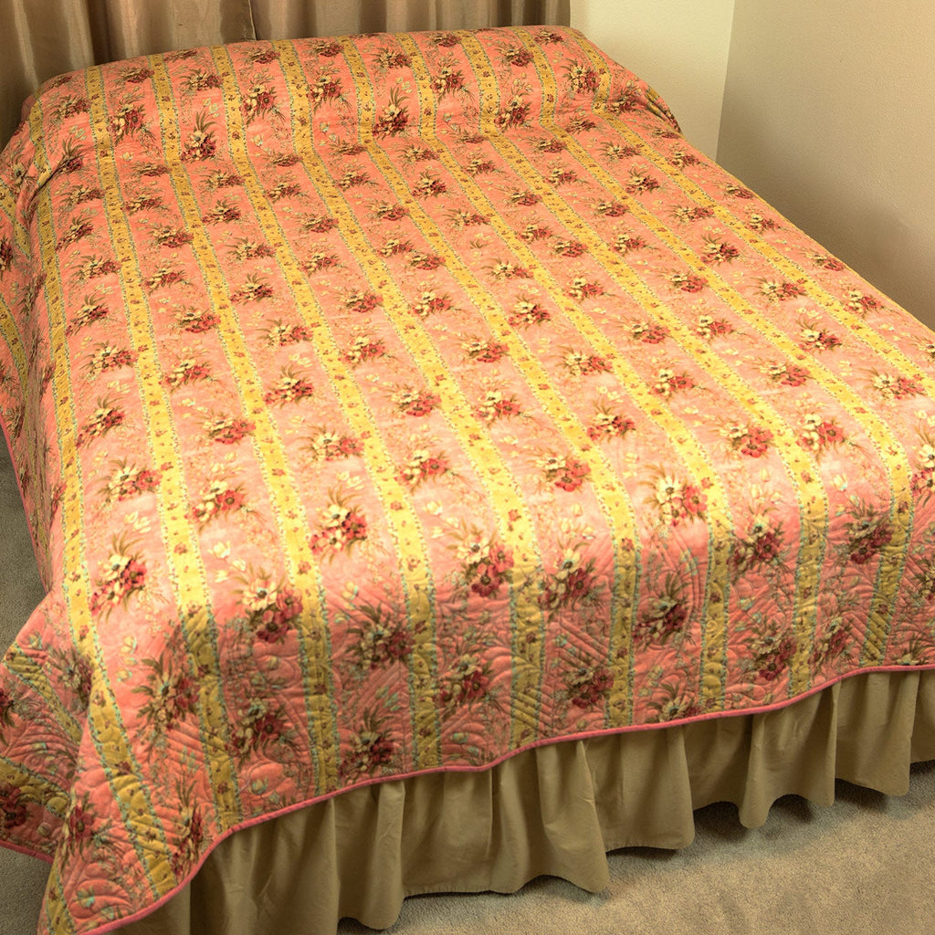 A pink quilted bedspread with a yellow striped pattern and floral motifs, styled on a bed with a beige ruffled bed skirt in a bedroom setting.