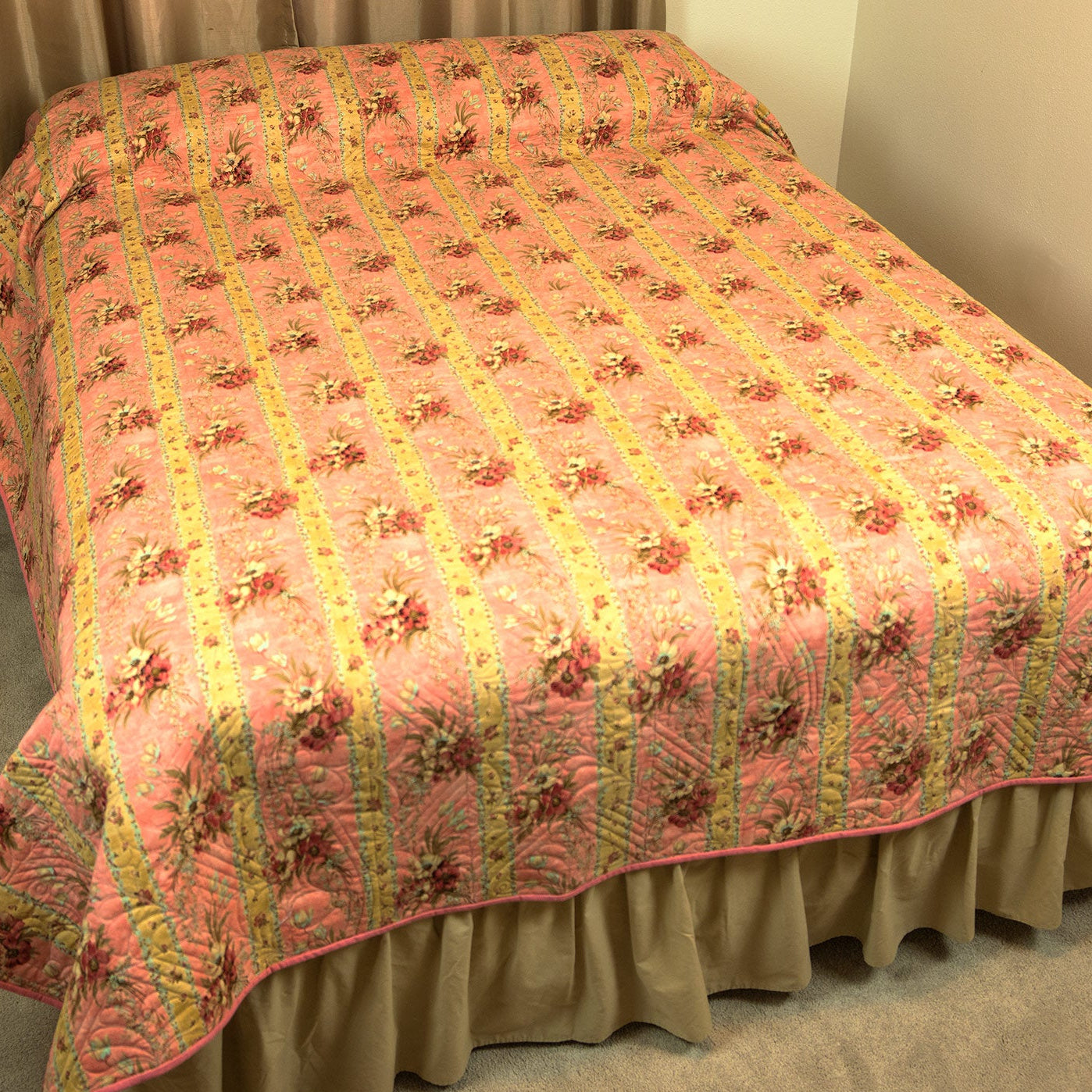 A pink quilted bedspread with a yellow striped pattern and floral motifs, styled on a bed with a beige ruffled bed skirt in a bedroom setting.