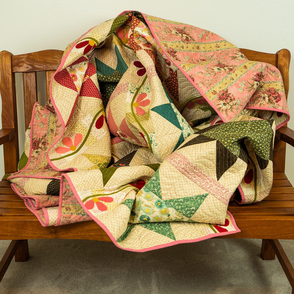 A multicolor patchwork quilt with pink, green, and beige floral and geometric patterns, draped over a wooden bench against a plain wall.