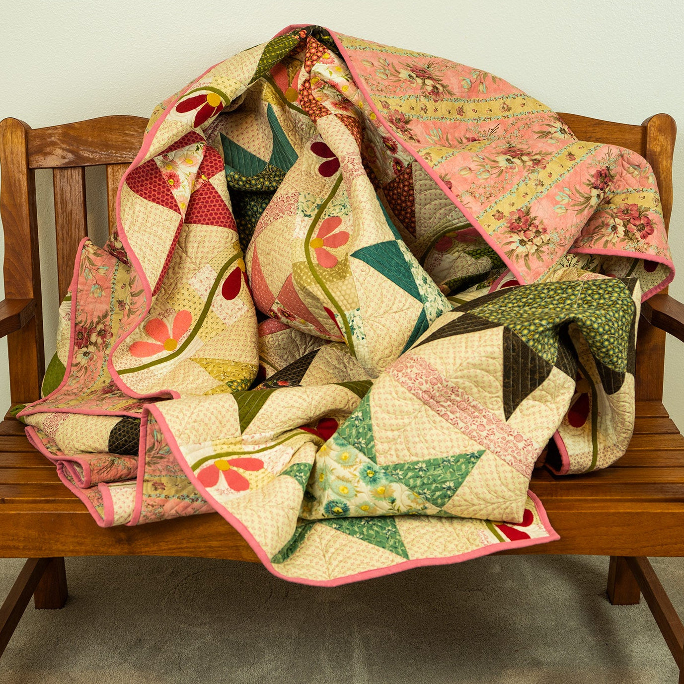 A multicolor patchwork quilt with pink, green, and beige floral and geometric patterns, draped over a wooden bench against a plain wall.