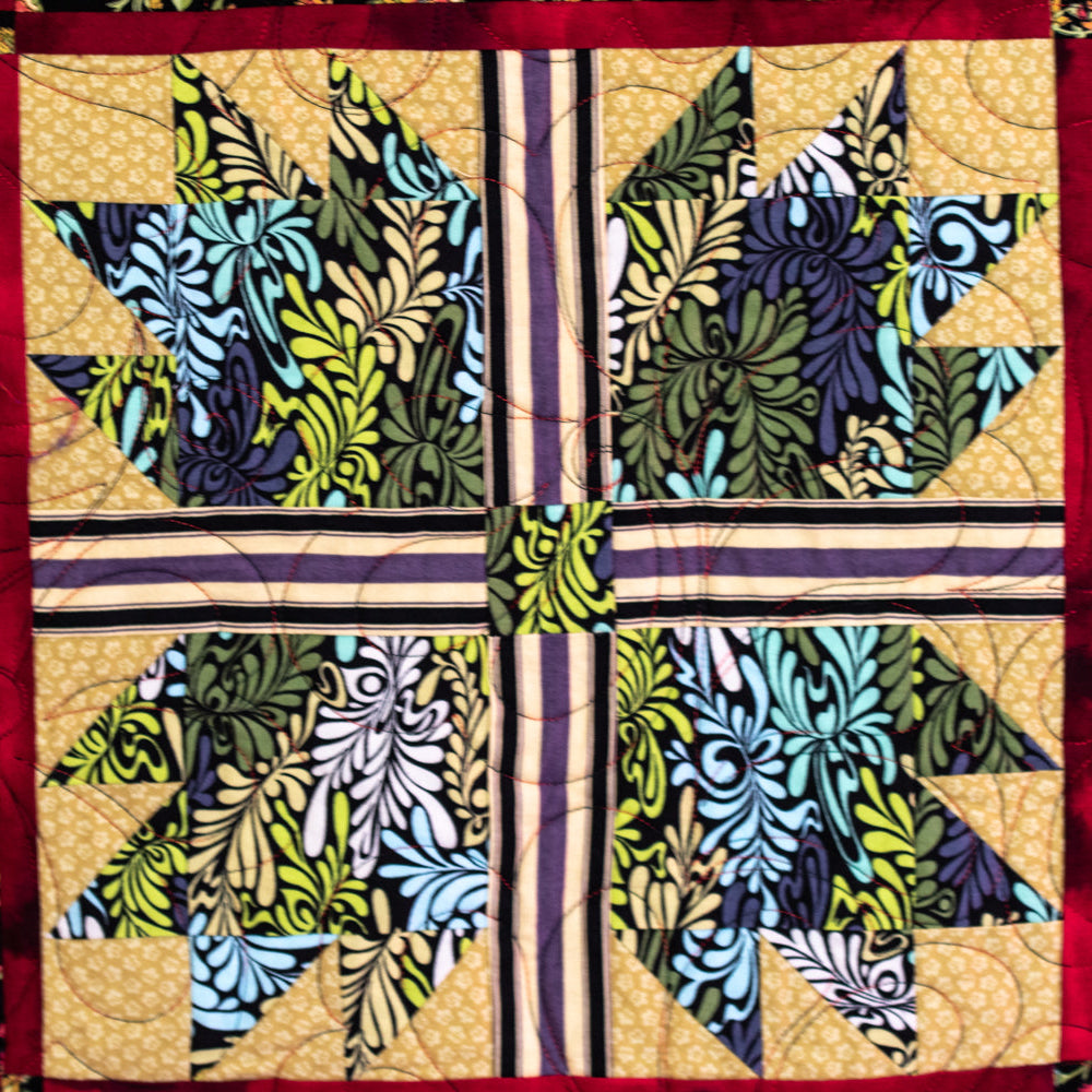 A close-up of a colorful quilted textile featuring a central geometric block design with floral and leafy patterns in blue, green, yellow, and purple, bordered by red and black floral fabric panels.