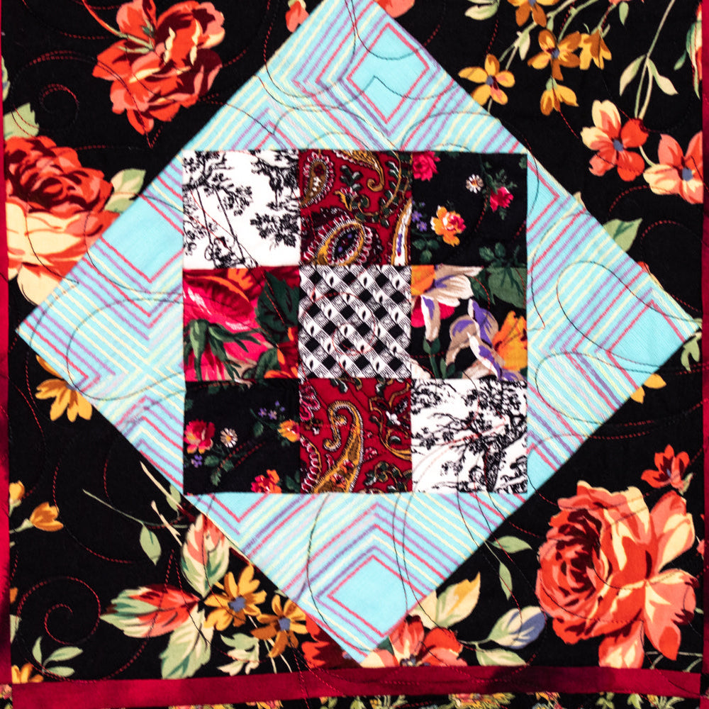 A close-up of a quilted textile featuring a central diamond patchwork of floral and paisley fabrics, surrounded by a light blue plaid section and a black floral border with red ribbon-like trim.