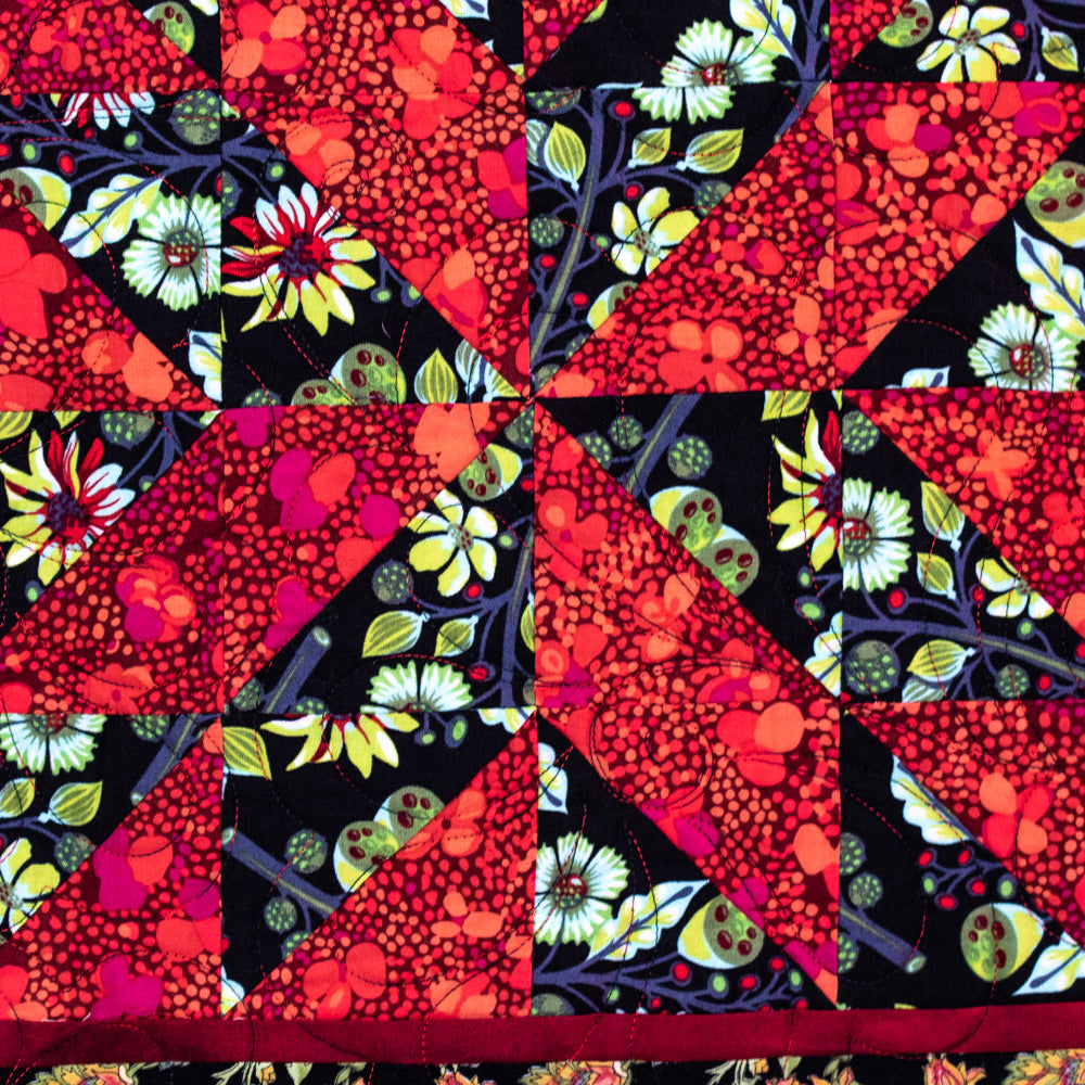 A close-up of a quilted textile featuring a geometric starburst pattern made from red floral and berry-print triangles over black backgrounds with white, yellow, and green floral motifs.