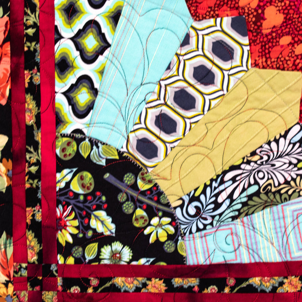 A close-up of a colorful quilted fabric panel with multiple patterned fabric sections arranged in a fan shape, bordered by a red stitched border. The fabrics include floral, geometric, and abstract prints in red, black, yellow, blue, and white.