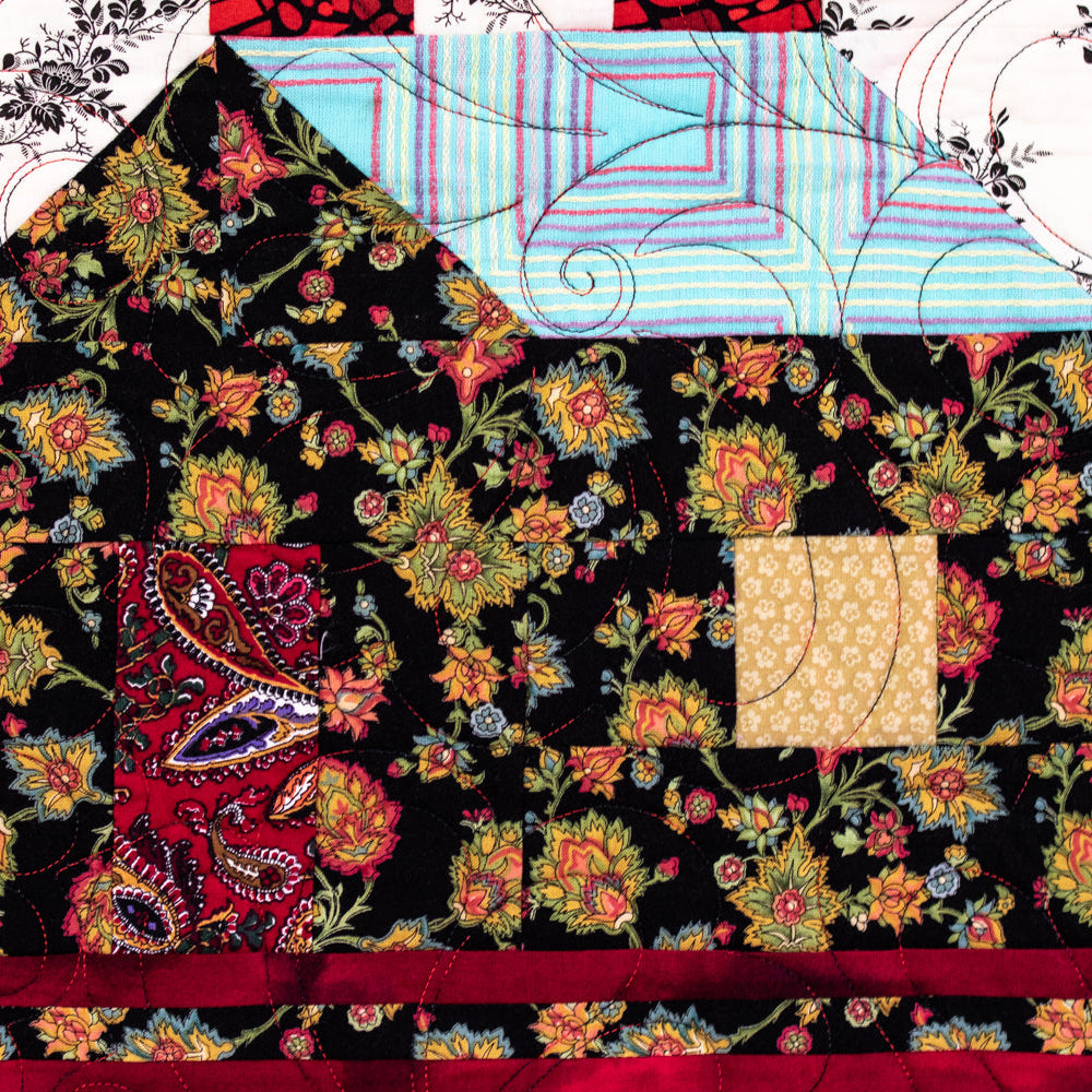 A close-up of a quilted patchwork design shaped like a house, featuring a black floral front, a light blue striped roof, and red borders.