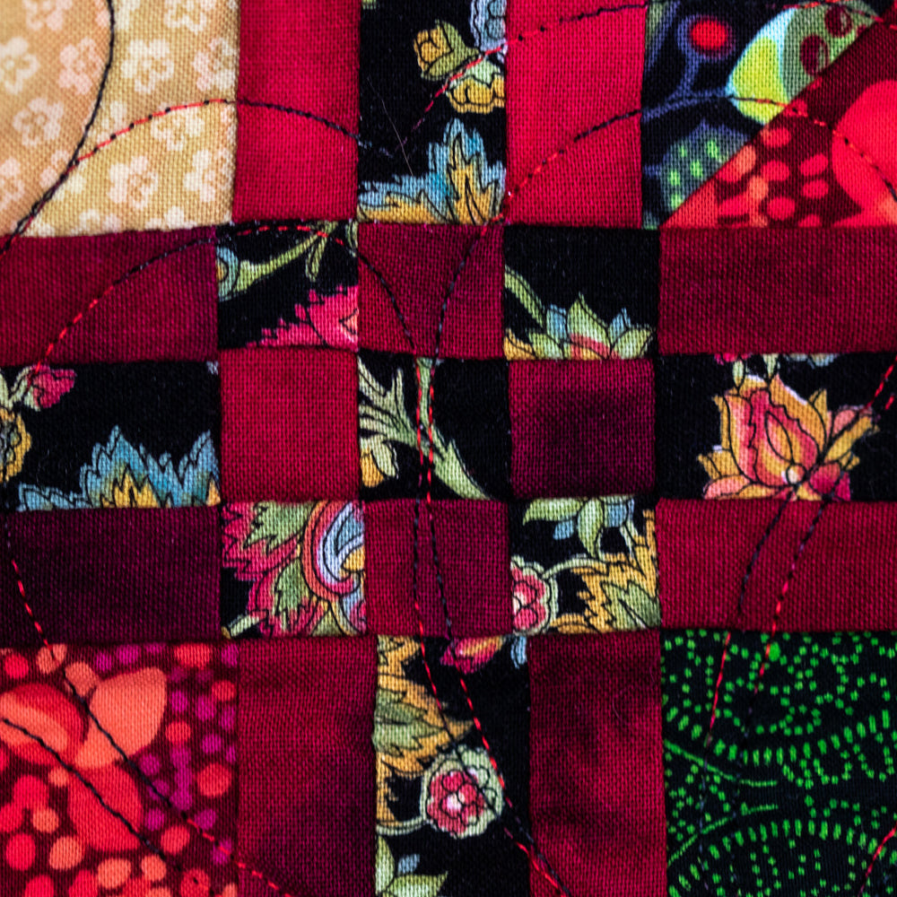 A close-up of a colorful quilted fabric block pattern featuring red, black, floral, and green patterned textile squares with visible running quilting stitches.