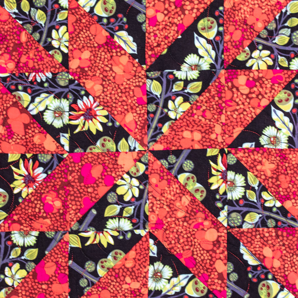 A close-up of a quilted textile featuring a geometric starburst pattern made from red-orange floral and dotted fabric triangles, set against black floral backgrounds with red borders.