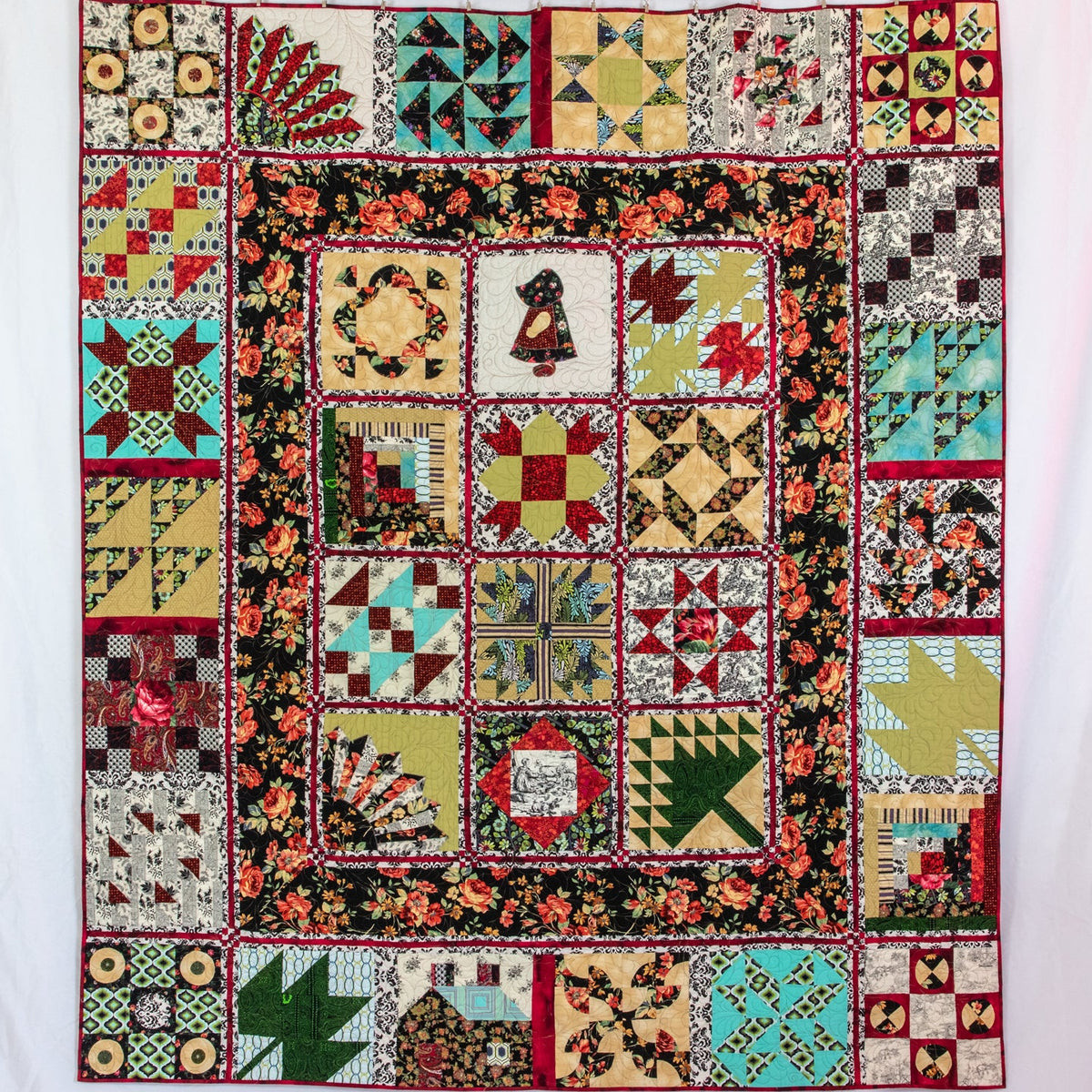 A vintage-style patchwork quilt hanging flat against a white backdrop. The quilt features a central floral border and a multicolor patchwork design with geometric and floral blocks in red, green, blue, yellow, black, and beige tones.
