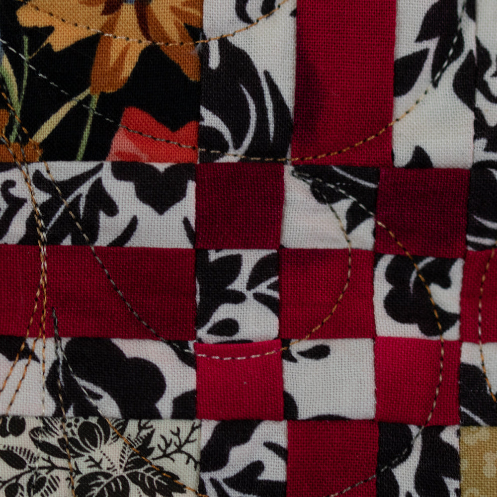 A close-up of a patchwork quilt showing red, black, and white patterned fabric blocks with floral and damask designs, stitched together with visible running stitch quilting.
