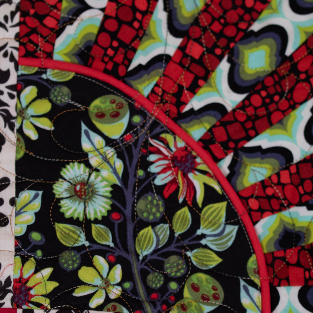 Close-up of a colorful quilted fabric with a floral pattern on a black background and radiating red, green, black, and white geometric stripes.