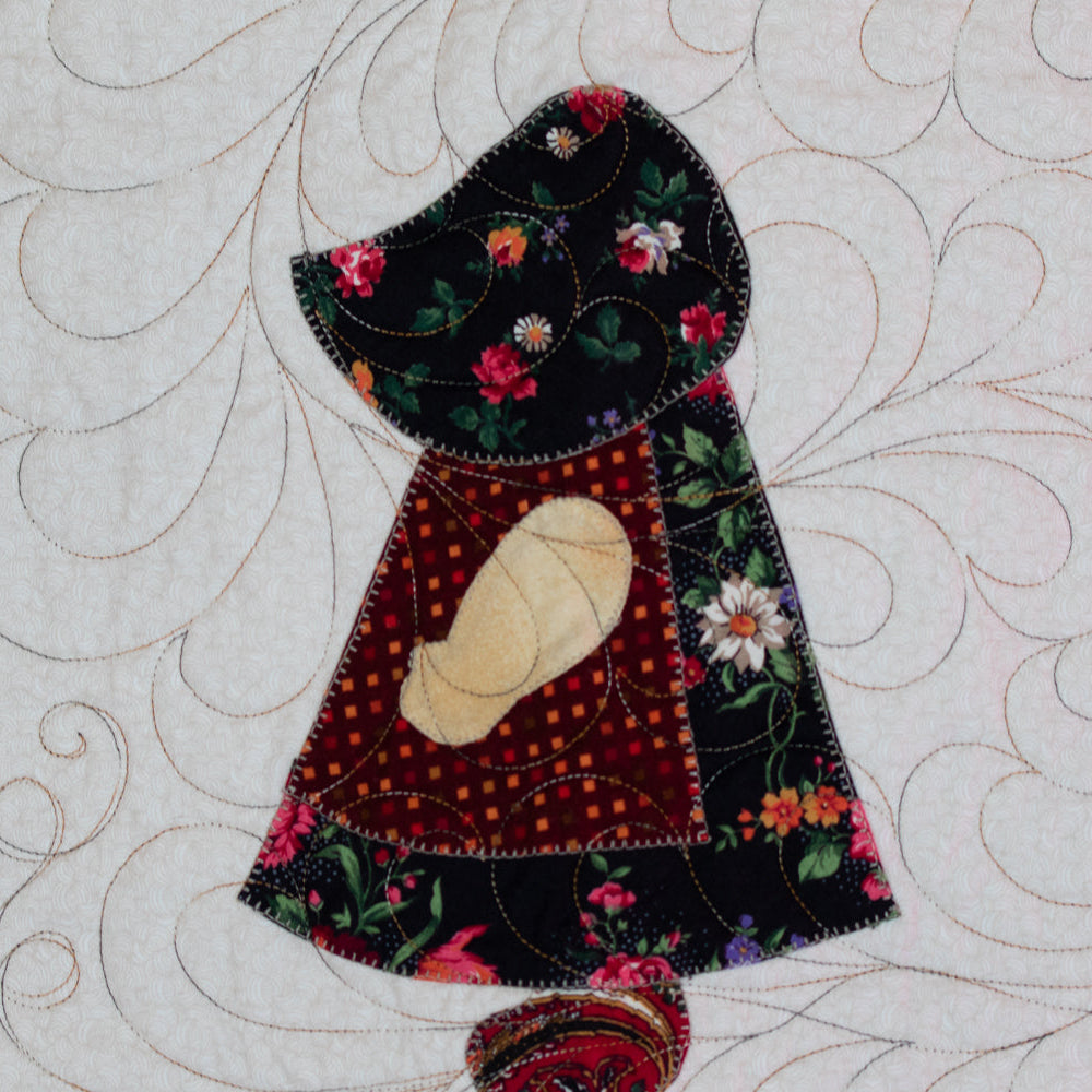 A close-up of a quilted textile featuring a stylized cloaked figure with floral fabric applique, a cream arm, and swirling stitched patterns on an off-white background.
