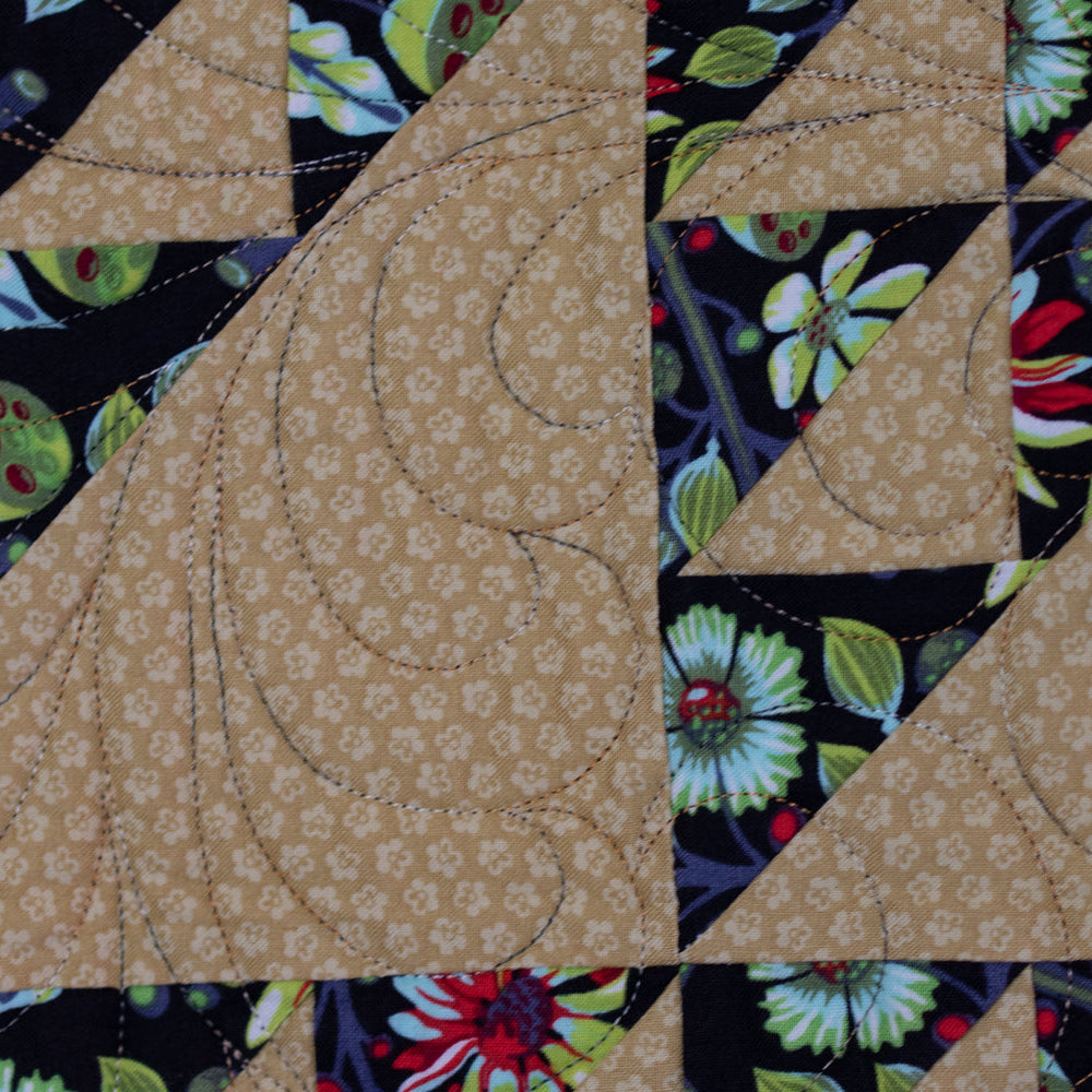 Close-up of a quilted textile with large tan geometric blocks bordered by dark navy floral and botanical fabric panels.