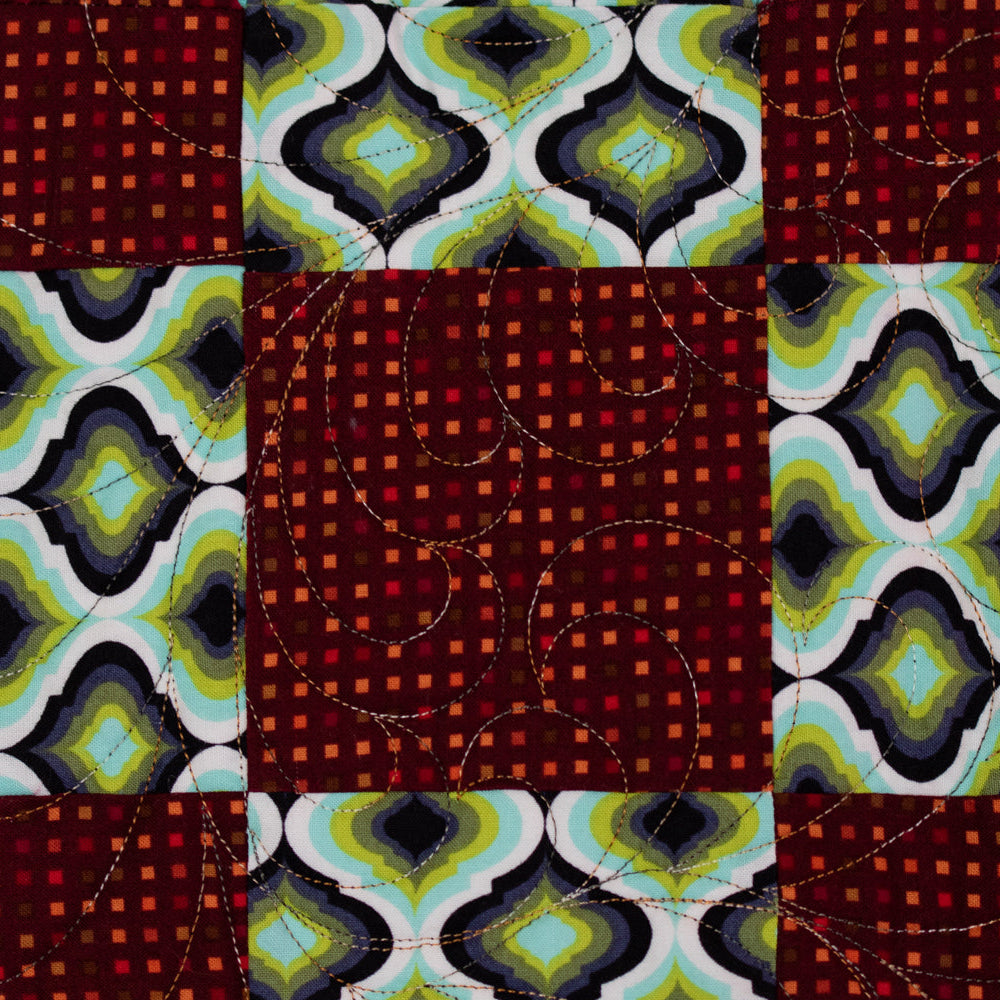 A close-up of a quilted textile featuring a geometric patchwork design with alternating blocks of dark red, black, green, white, and turquoise patterns, including wavy quilting stitches.