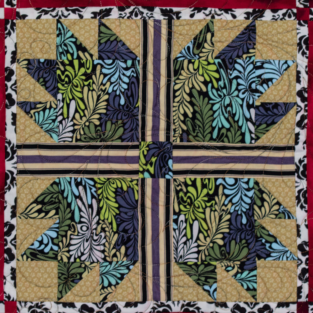 A close-up of a quilted textile featuring a central cross-block design with floral and leafy patterns in teal, green, navy, and beige, bordered by red and black-and-white patterned fabric.