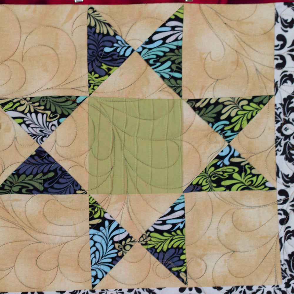 A close-up of a quilted fabric panel featuring a geometric starburst pattern. The center is a sage green square, surrounded by beige diamond sections with swirling stitched lines, flanked by red, black-and-white damask, and floral patterned fabric sections.
