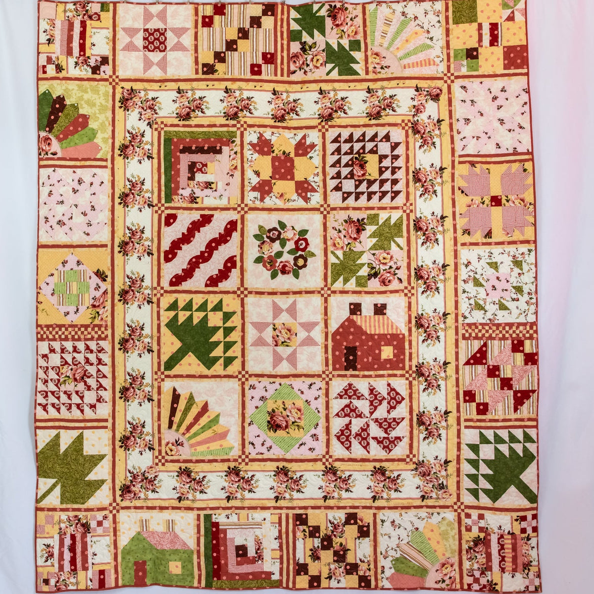 A square quilted patchwork blanket featuring a rustic country design with various geometric and floral blocks in muted tones of red, green, beige, and brown, hung flat against a white background.