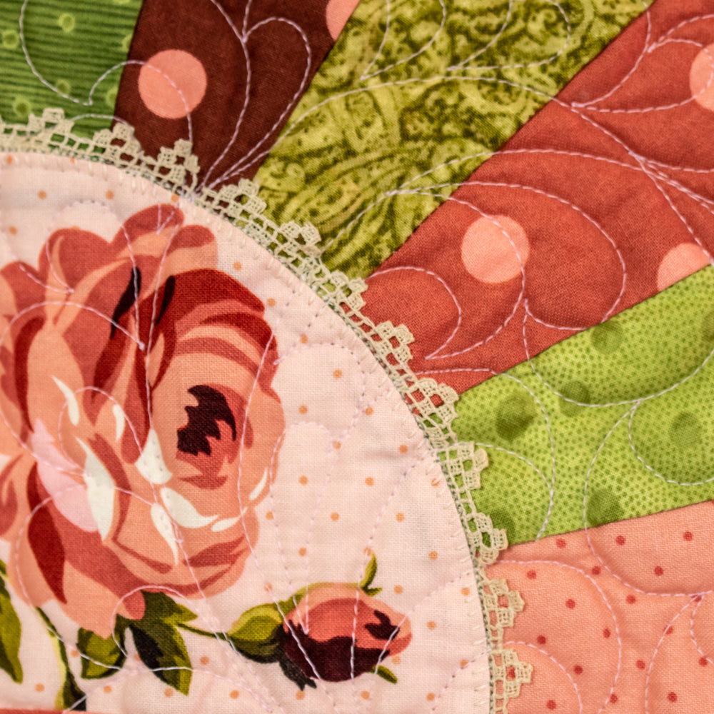Close-up of a quilted fabric panel with a floral rose motif, lace trim, and curved stitched sections in pink, green, rust, and peach tones.