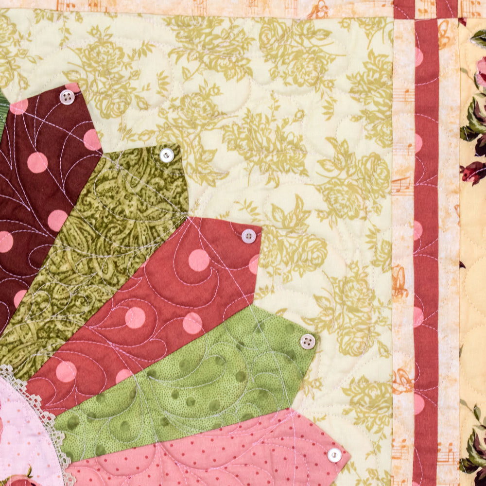 Close-up of a quilted fabric panel with layered fan-shaped patches in pink, green, and burgundy on a cream floral background, bordered by pink roses and floral print.