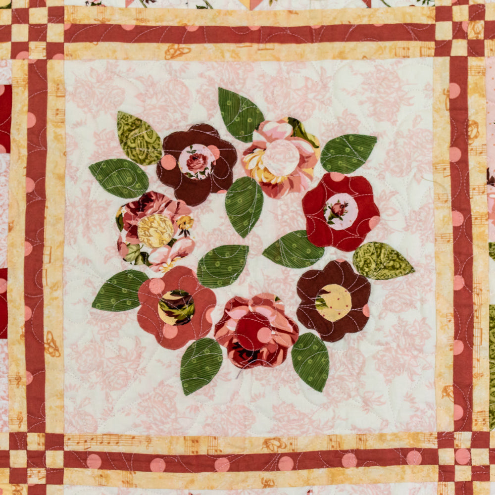 A close-up of a quilted fabric panel featuring a floral wreath design with red, pink, and dark brown flowers, green leaves, and a cream background, bordered by red and gold checkered stripes.