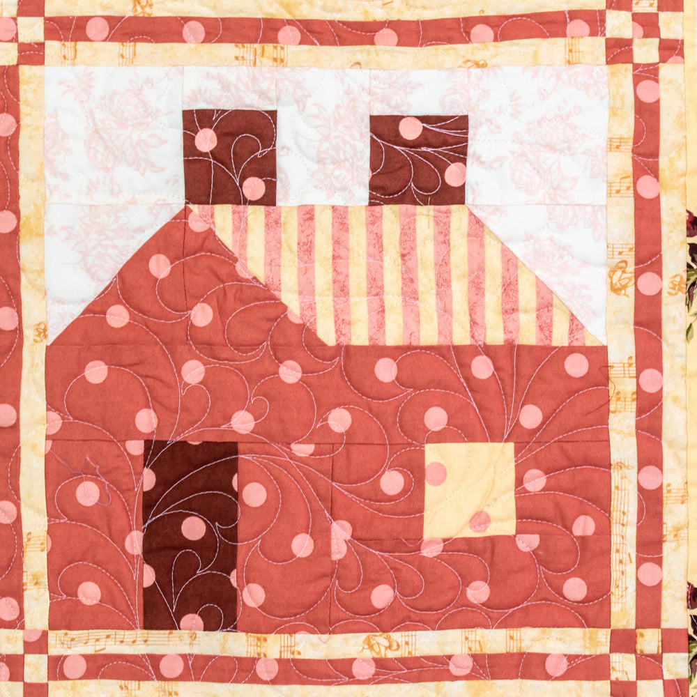 A close-up of a quilted textile featuring a stylized house motif in terracotta, pink, cream, and brown, bordered by floral fabric with pink roses and yellow accents.