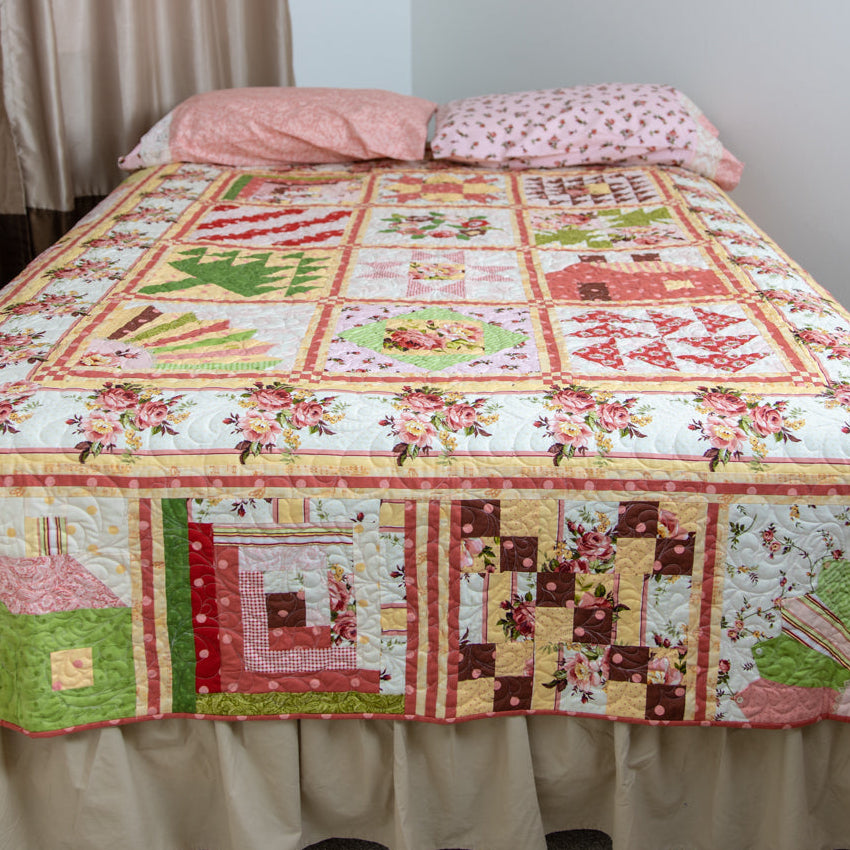 A patchwork quilted bedspread with a rustic cabin and floral pattern, styled on a bed with a beige ruffled skirt and pink pillows in a bedroom setting.