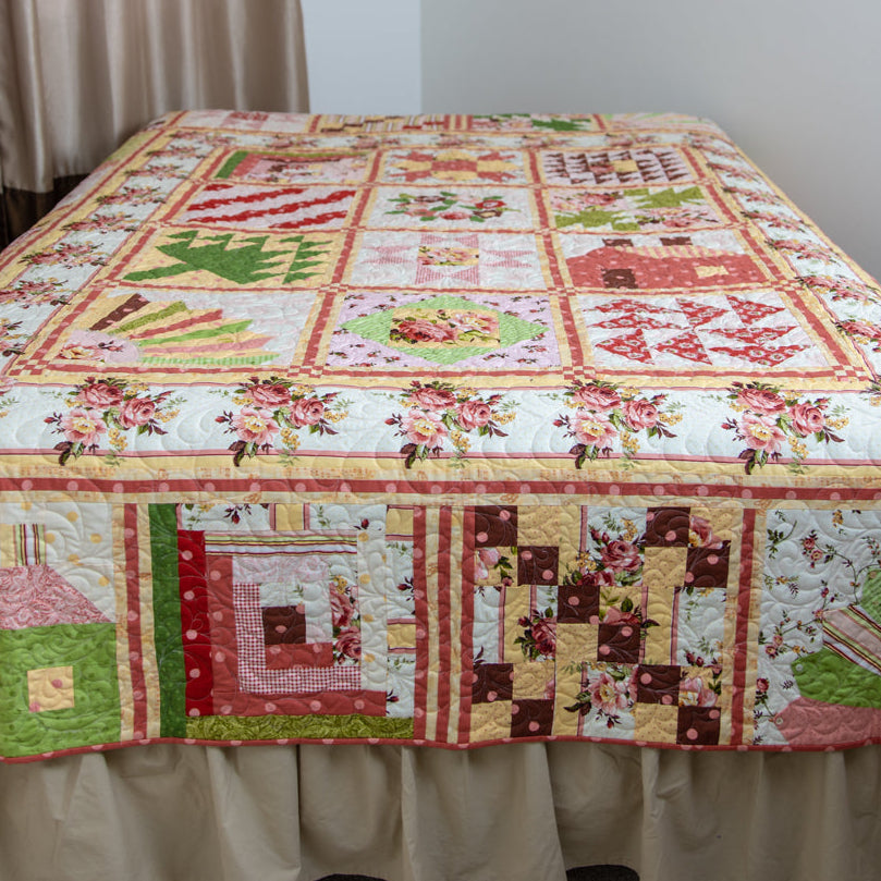 A patchwork quilted bedspread with a rustic country theme, featuring floral patterns and small house motifs, draped over a bed with a beige ruffled skirt in a bedroom setting.