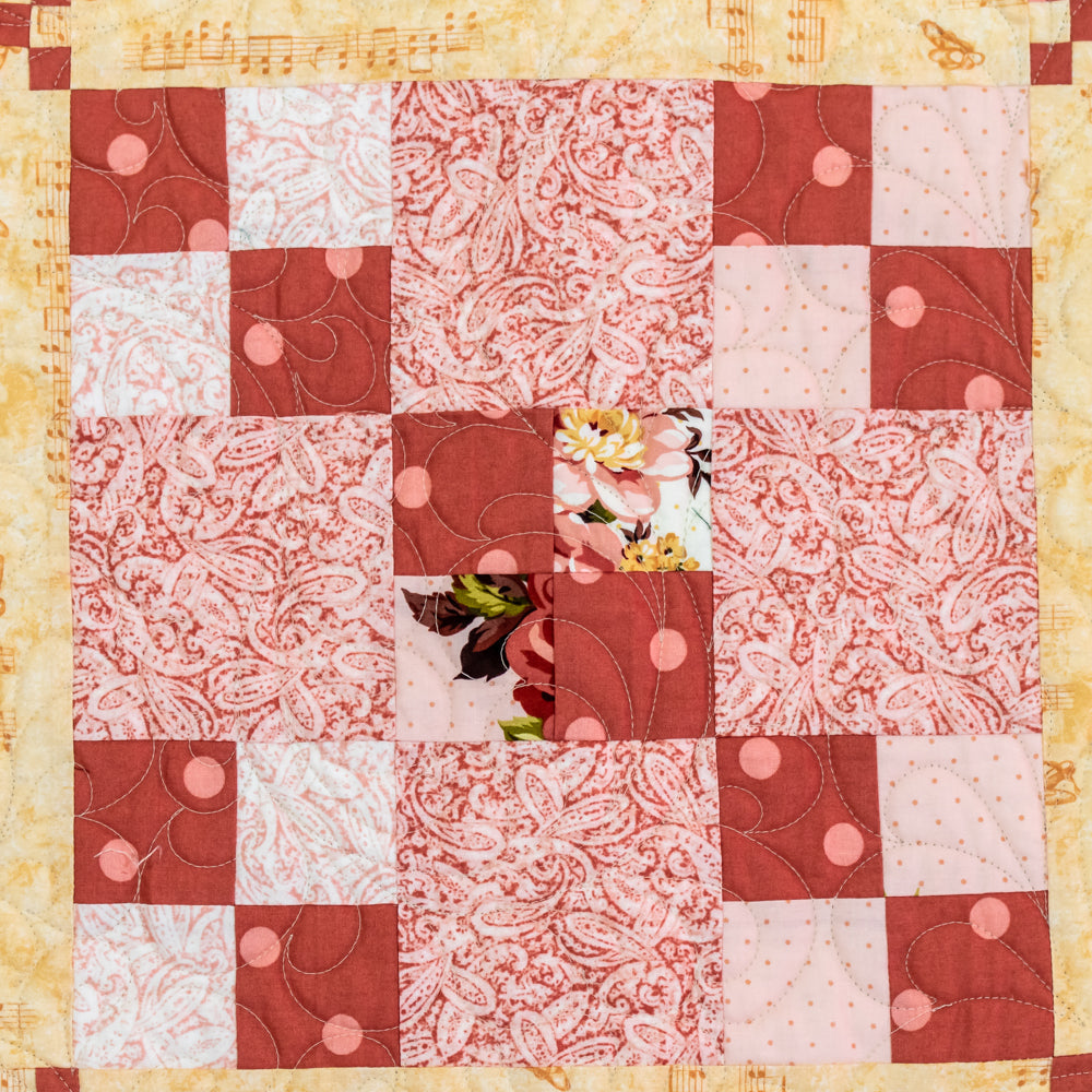 Quilt block with a square checked pattern made up of floral and polka dot prints in pinks and neutrals. There is also a border in a neutral music note print.