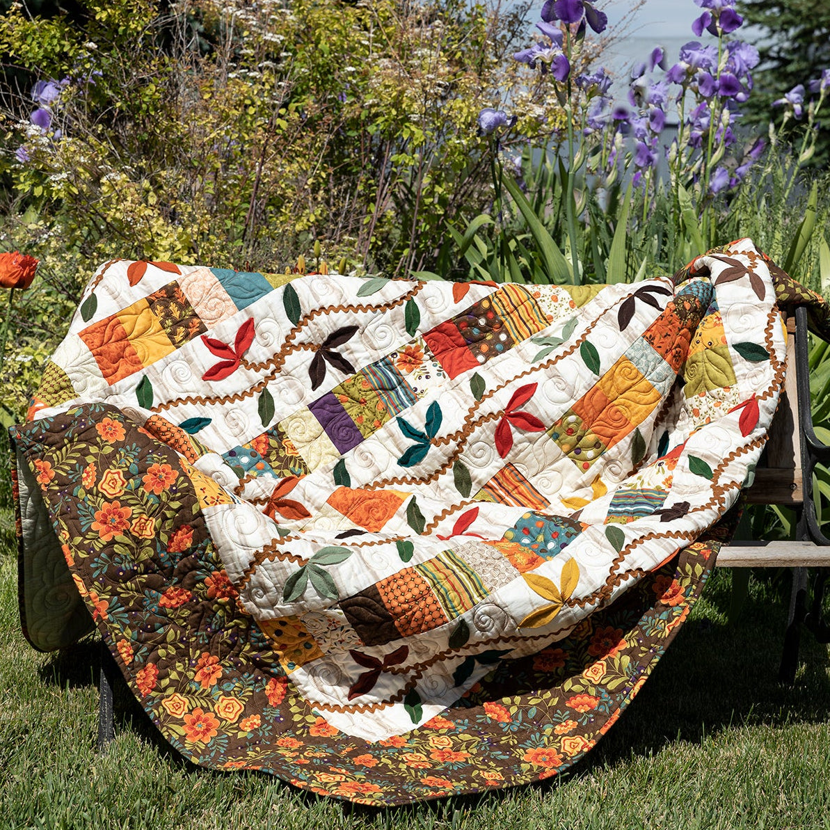 A colorful patchwork quilt draped over a black metal garden bench in a grassy backyard with purple flowers and green foliage in the background.