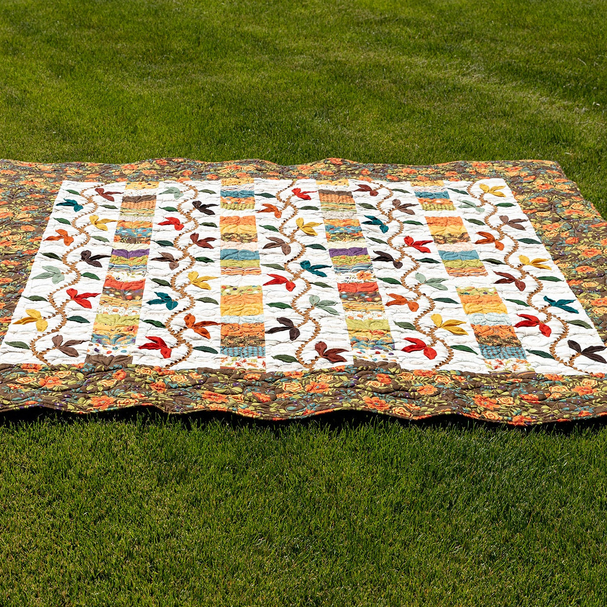 A colorful patchwork quilt with a floral border and butterfly motifs, laid flat on a green grass lawn.